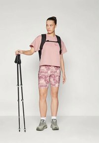 Rozā īsām piedurknēm blūze, rakstainas rozā un baltas riteņbraukšanas šortas, pelēki pārgājienu apavi un melna mugursoma; rokās turēt trekinga nūjas.