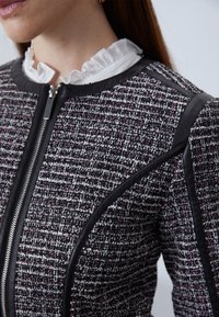 Black and white tweed jacket featuring a zip closure, leather accents, and a ruffled white collar, with a structured and fitted design.