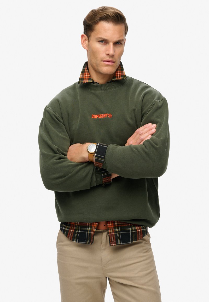 Groene fleece sweatshirt met oranje logo, gedragen over een zwart-rood geruite shirt. Casual pasvorm met ribgebreide boorden. Tan broek.