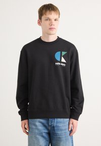 Crna dukserica s okruglim izrezom, s obojenim logotipom i tekstom na lijevoj strani prsnog dijela. Glatka tkanina, opušteni kroj i dugi rukavi.