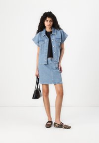 Jachetă din denim albastru deschis cu mâneci scurte, fustă din denim asortată, top negru și balerini cu model, completată cu o mică geantă de mână neagră.