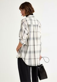 Kvinna med kort brunt hår iförd en lös vit och svart rutad skjorta och svarta byxor, som håller en svart handväska vid sin sida.