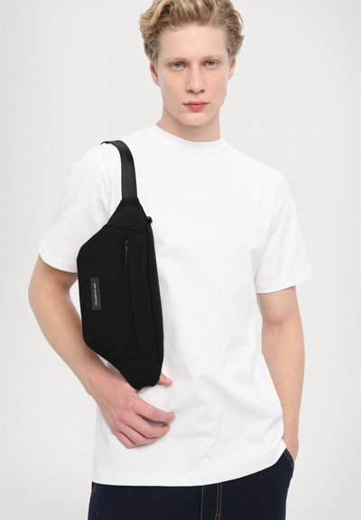 Svart stoff sling-veske med glidelåslomme, båret over en hvit t-skjorte. Har en teksturert overflate og justerbar stropp.