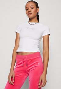 Vit kortärmad cropped topp med "Juicy" i silver rhinestones, matchad med rosa sammetsoverall med dragsko och dekorativ detalj.