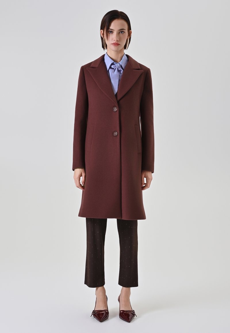 Cappotto di lana borgogna con un design sartoriale, due bottoni e tasche laterali, abbinato a pantaloni scuri a fantasia e scarpe a punta.