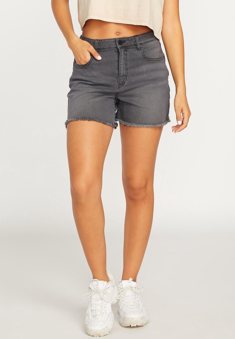 Femme portant un short en jean gris à ourlets effilochés, un crop top beige, des baskets blanches, la main gauche dans la poche, debout devant un fond uni.