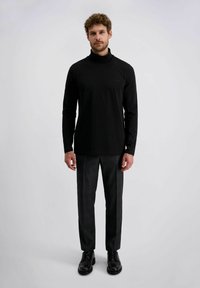 Ein Mann trägt ein schwarzes Rollkragen-shirt, dunkelgraue Anzughose und schwarze Anzugschuhe und steht vor einem schlichten hellgrauen Hintergrund.