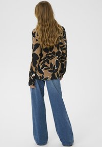 Vrouw met lang golvend haar, gekleed in een losvallende bruin- en zwartgepatrooneerde shirt en wijde blauwe jeans, staand tegen een witte achtergrond.
