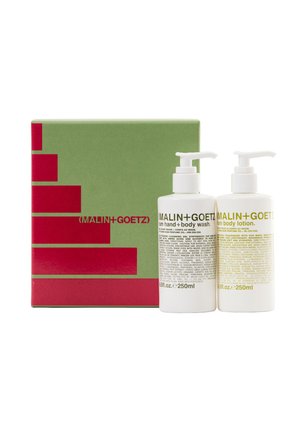 Weiße Pumpspender-Flaschen mit Handseife und Körperlotion, mit gelbem Etikett, vor einer grünen Geschenkbox mit rotem geometrischem Muster.