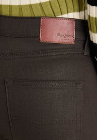 Tummanruskeat farkut, joissa on pehmeä tekstiili ja takatasku näkyvissä. Niissä on myös tarra-merkki, jossa lukee "Pepe Jeans London".