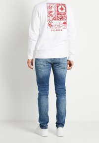 Bílý sweatshirt s dlouhým rukávem a červenými grafickými potisky na zadní straně, kombinovaný s modrými džíny v úzkém střihu a s lehkým práním.