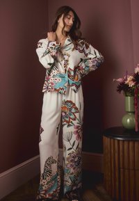 Woman wearing cream-colored floral and zebra print pajama set stands in a corner next to a round wooden table with a green vase of flowers.