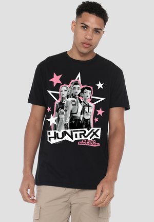 Young man wearing a black T-shirt with three animated female characters, pink and white stars, and "HUNTR X K-POP DEMON HUNTERS" text.