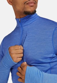 Camiseta deportiva azul con cremallera y cuello tipo mock. Cuenta con mangas largas, agujeros para los pulgares y una textura suave con rayas tonales sutiles.