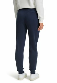 Navyblauwe sweatpants met een getailleerde pasvorm, geribde boorden en details op de achterzak. Gepaard met witte sneakers op een neutrale achtergrond.