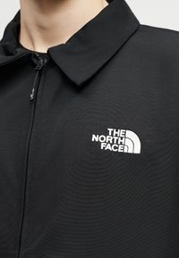 Κοντινό πλάνο ενός ατόμου που φοράει μαύρο μπουφάν με φερμουάρ, γιακά και λευκό λογότυπο "The North Face" στο στήθος.