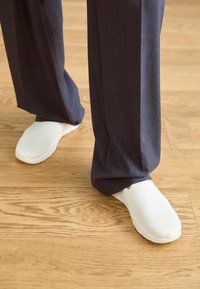 Vita slip-on-skor med en texturerad ovandel, tillsammans med mörkgrå vida byxor, mot en träggolv.