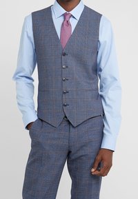 Man wearing a blue checkered suit vest and matching pants, light blue shirt, and textured pink tie, standing with hands partially in pockets.
