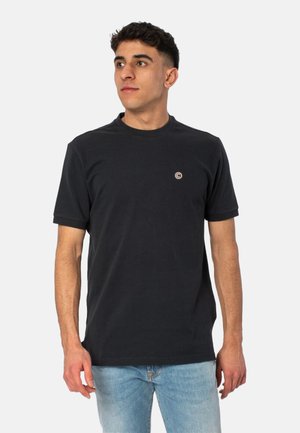 T-shirt nera in cotone con scollatura rotonda e maniche corte, caratterizzata da un piccolo logo circolare sul petto. Indossata con jeans blu chiaro.
