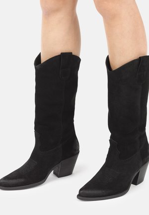 Stivali cowboy in camoscio nero fino a metà polpaccio con punte leggermente appuntite e tacchi impilati, indossati su gambe nude contro uno sfondo neutro.