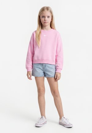 Ung pige står med fronten fremad iført en lyserød sweatshirt, denimshorts og hvide sneakers mod en helt hvid baggrund.