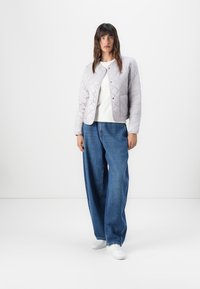 Light gray quilted jacket with a round neckline, snap buttons, and two side pockets, paired with loose-fit blue jeans and white sneakers.
