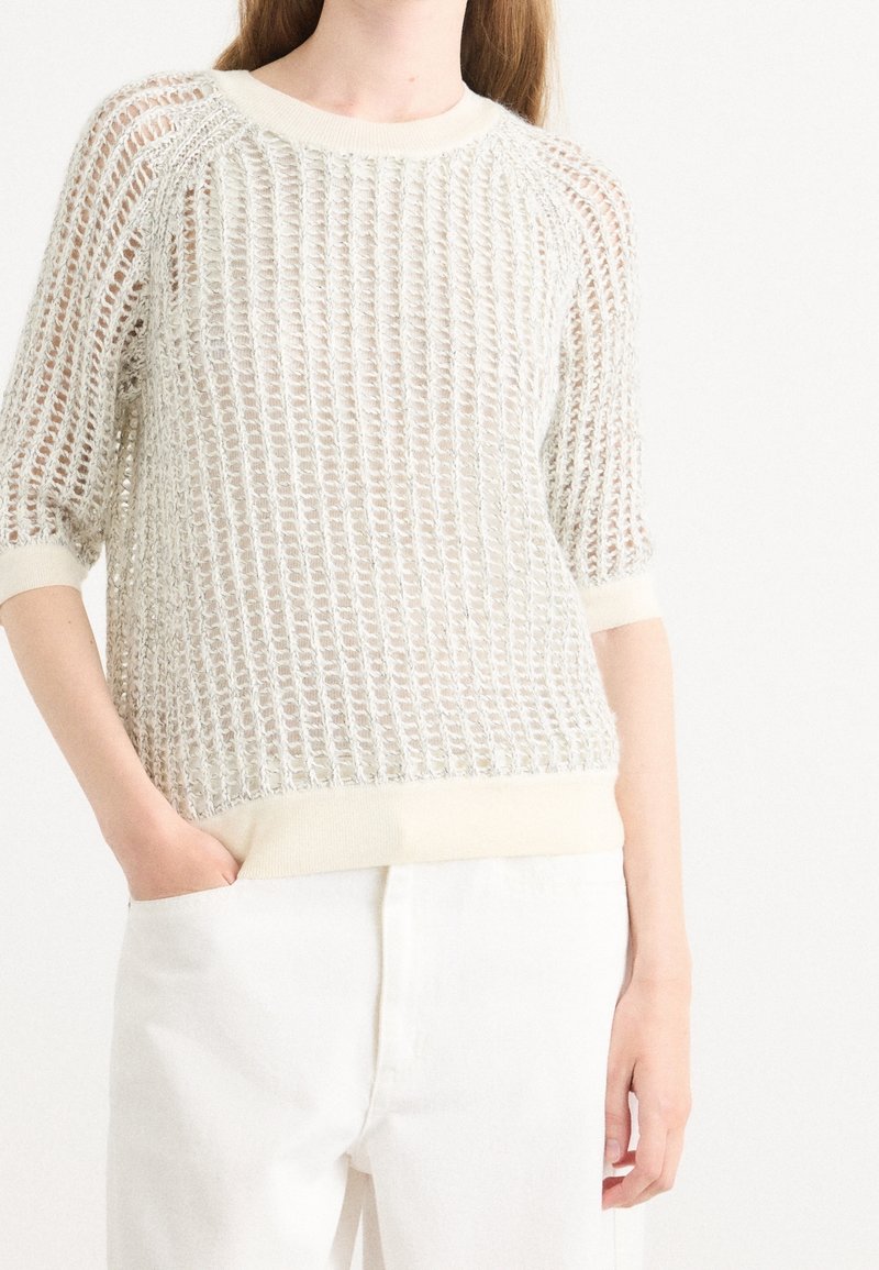 Personne portant un pull court manches en maille ample de couleur crème sur un haut blanc, associé à un pantalon blanc taille haute, main dans la poche.