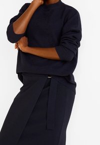 Ciemnoniebieski sweter z wysokim kołnierzem i opadającymi ramionami, w zestawie z dopasowaną spódnicą ołówkową z bocznym wiązaniem. Miękka, teksturowana tkanina.