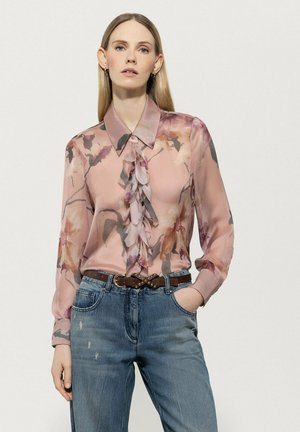 Bluse mit floralem Muster, aus sheer pinkem Stoff, mit Kragen und gerafftem Vorderteil. Kombiniert mit hellblauen, ausgewaschenen Jeans und einem braunen Gürtel.