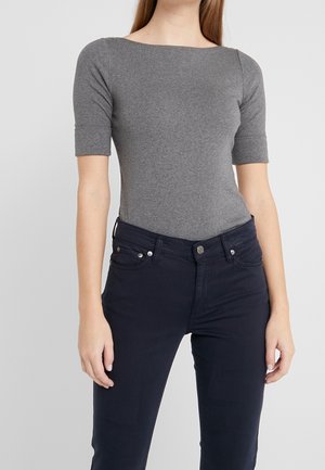 Kobieta w dopasowanym szarym topie z krótkim rękawem i ciemnoniebieskich dżinsach, stojąca na tle jednolitego jasnoszarego tła.