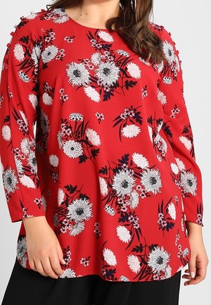 Frau trägt eine rote Langarmbluse mit weißen und schwarzen Blumenmustern, kombiniert mit schwarzen Hosen.