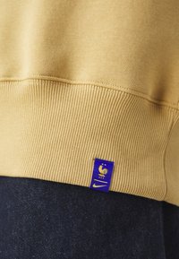 Beżowa, prążkowana sweatshirt z dolnym wykończeniem i małą, niebieską metką z złotym kogutem, dwoma gwiazdkami, literami FFF oraz żółtym logo Nike w kształcie łyżwy.