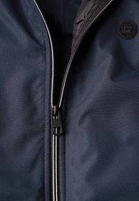 Detailní záběr na částečně rozepnutou tmavě modrou bundu se stříbrnými zuby zipu, černým poutkem na zip a černým kulatým tlačítkem s geometrickým vzorem.