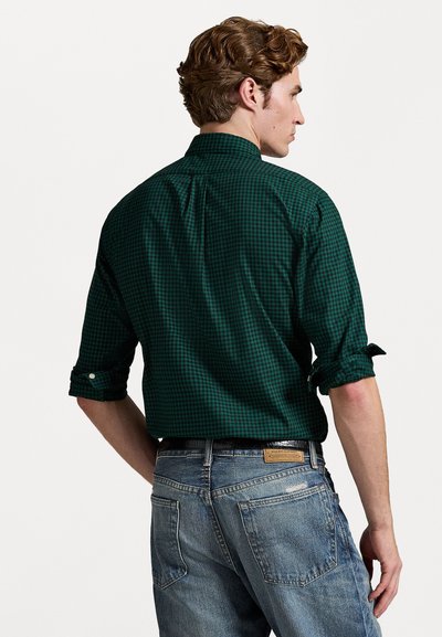 Mørk grønn rutete skjorte med oppbrettede ermer og knapper på mansjettene, sammen med lyseblå denim jeans, som har et merke på baklommen.