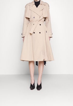Manteau trench-coat beige à double boutonnage avec ceinture nouée, col large et boutons, porté avec des chaussures noires à bouts pointus sur un fond uni.
