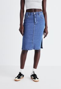 Vidēja zila džinsu zīda svārki ar priekšu šķēlumu un piecām kabatām, ar rāvējslēdzēju un metāla pogu aizdari, valkāti kopā ar melniem sneakeriem.