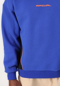 Felpa blu con orlo e polsini a coste, dotata di un logo arancione sul petto e dettagli neri ai lati. Tessuto morbido e strutturato.
