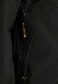 Schwarze Stoffjacke mit einer Reißverschlusstasche. Merkmale eines sichtbaren Markenaufnähers "PME LEGEND" in Gold am Reißverschluss. Glatte Textur.