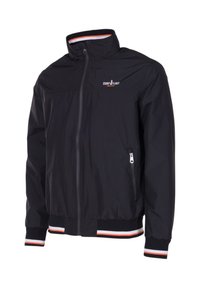 Zwarte jas met ritssluiting, hoge kraag, gestreepte boorden en taille en een klein logo op de linkerborst met de tekst "Seaside Legacy Beachwear."