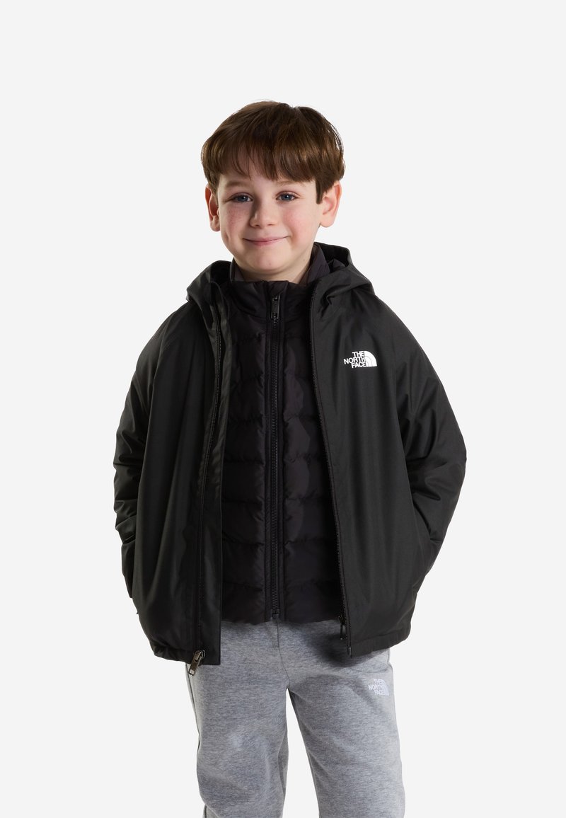 Αγόρι που φοράει μαύρο μπουφάν North Face και γκρι φόρμες, στέκεται με τα χέρια στις τσέπες του μπουφάν, χαμογελώντας στην κάμερα.