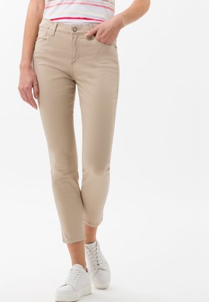 Pantalones slim-fit beige con una textura suave, que presentan un cierre de botón, bolsillos frontales y puños enrollados, combinados con zapatillas blancas.