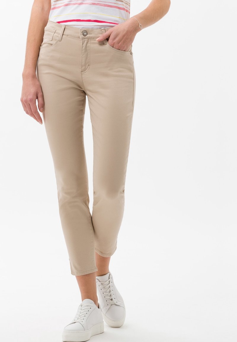 Pantalones slim-fit beige con una textura suave, que presentan un cierre de botón, bolsillos frontales y puños enrollados, combinados con zapatillas blancas.