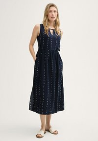 Marineblaues ärmelloses Kleid mit Muster aus weißen Punkten, taillierter Schnitt, seitlichen Taschen und einem Bindeband am Ausschnitt. Glattes, leichtes Material.