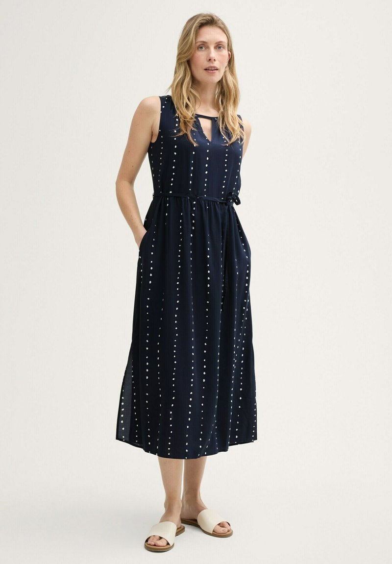 Robe sans manches bleu marine avec motif à pois blancs, taille cintrée, poches latérales et un détail de nouage au niveau du col. Tissu lisse et léger.