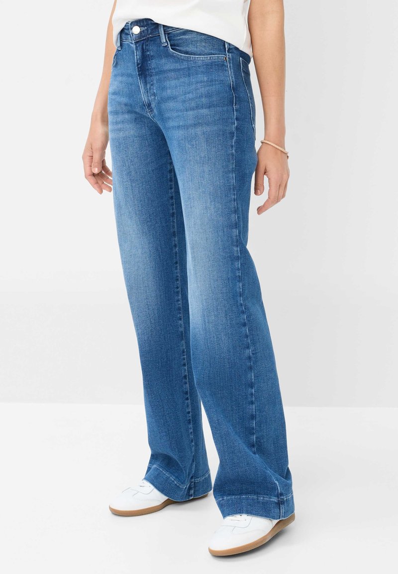 Personne portant un jean droit bleu et des baskets blanches, debout devant un fond clair uni.