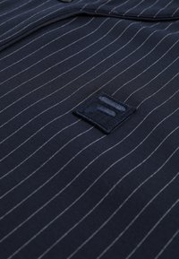 Marineblaue Nadelstreifen-Stoff mit einem kleinen, bestickten quadratischen Aufnäher, der drei horizontale, erhabene Streifen zeigt.