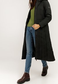 Kobieta idąca w niebieskich dżinsach, ciemnobrązowych butach, zielonym swetrze i długim czarnym pikowanym płaszczu z zamkiem błyskawicznym.