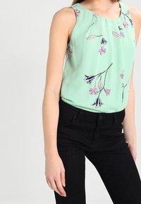 Blusa senza maniche verde menta con stampa floreale in rosa e nero. Tessuto liscio con scollo plissettato. Abbinata a pantaloni neri a righe verticali.