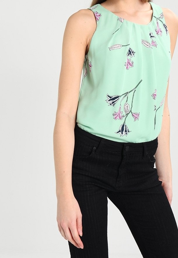 Blusa senza maniche verde menta con stampa floreale in rosa e nero. Tessuto liscio con scollo plissettato. Abbinata a pantaloni neri a righe verticali.
