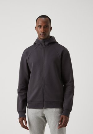 Felpa con zip di colore grigio scuro, dotata di cappuccio e maniche a raglan, con una texture morbida e polsini a coste. Indossata sopra pantaloni della tuta grigi chiari.